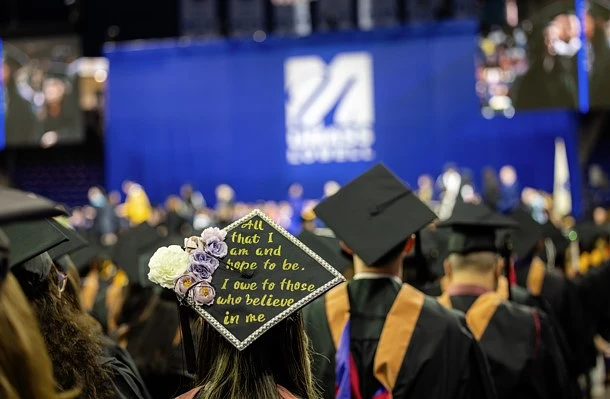 Graduates at a commencement ceremony stand facing a stage, with one decorated cap reading, "All that I am and hope to be, I owe to those who believe in me."