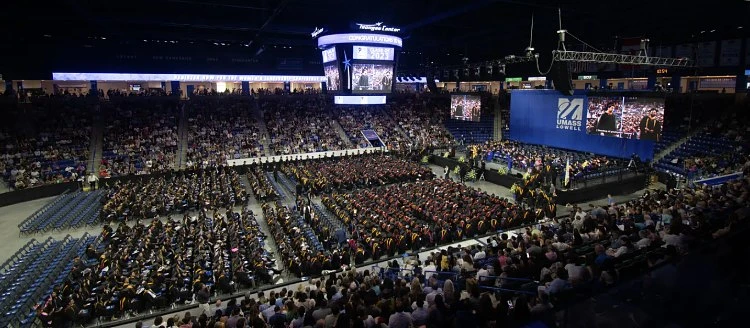 A large graduation ceremony is taking place in an indoor arena filled with seated graduates and spectators.